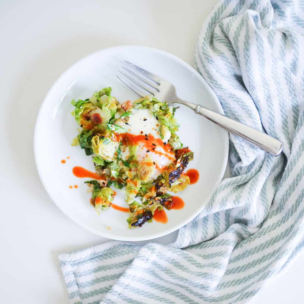 Whole30 Brussels Sprouts Hash topped with hot sauce on a white plate with a fork and towel on the side