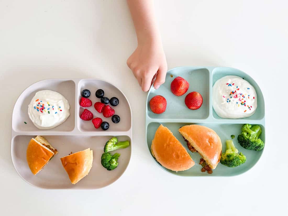 two divided plates with veggies, halved sandwiches, fresh fruit, veggies, and a treat