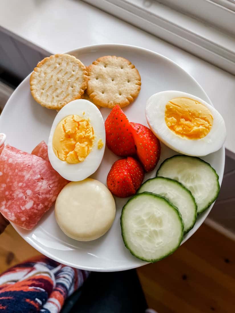a plate with crackers, hard boiled eggs, salami, a round piece of cheese, sliced strawberries, and cucumbers