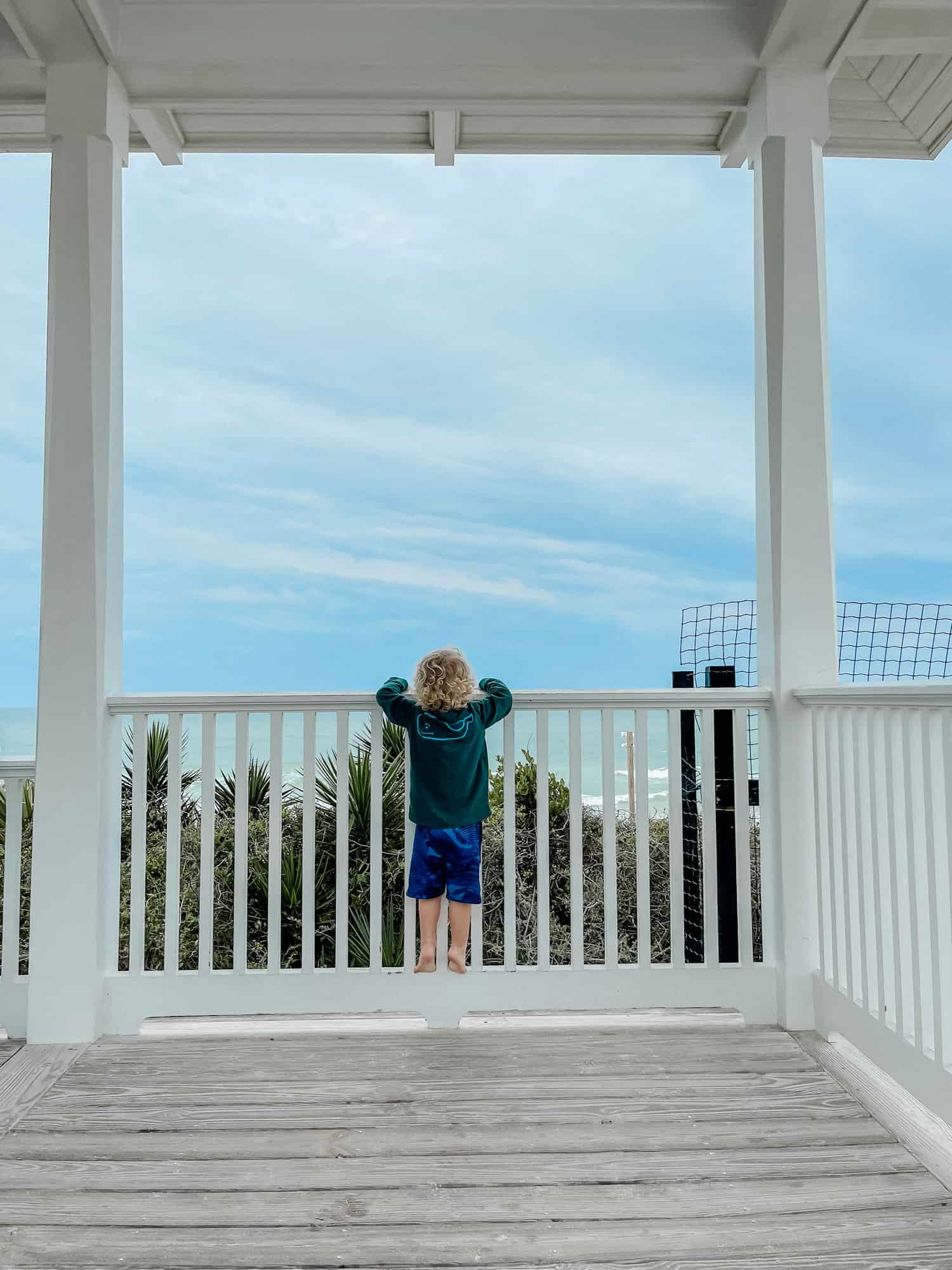 a little boy hanging off a porch railing looking at the beach 