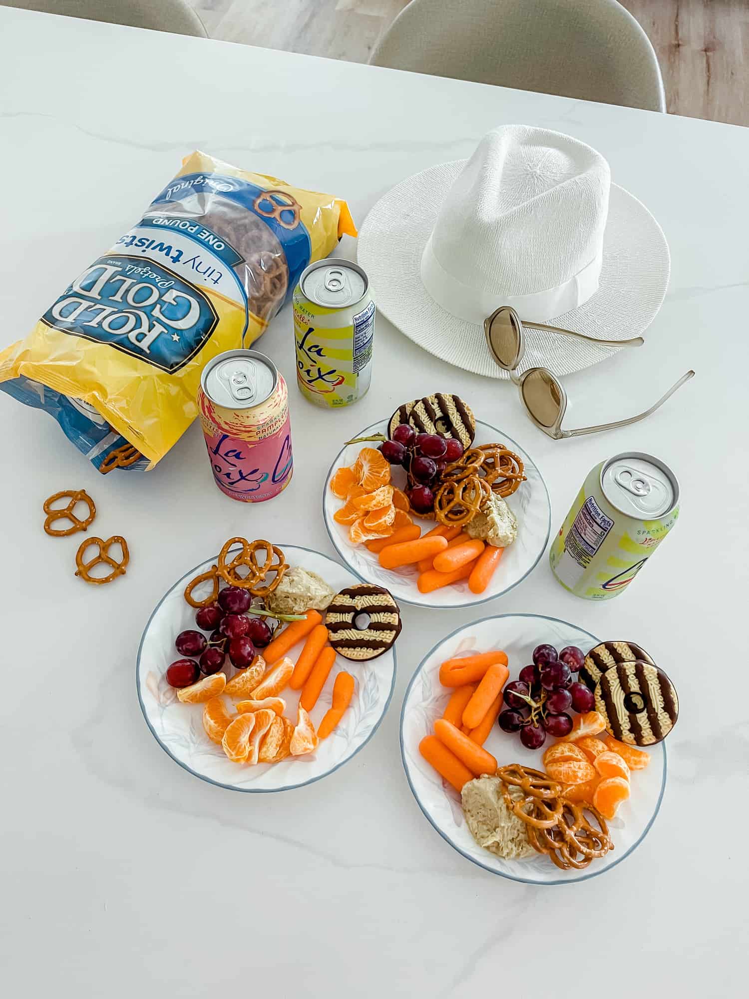 three plates filled with carrots, grapes, pretzels, orange slices, and cookies, a bag of pretzels, three la croix cans, a hat, and sunglasses