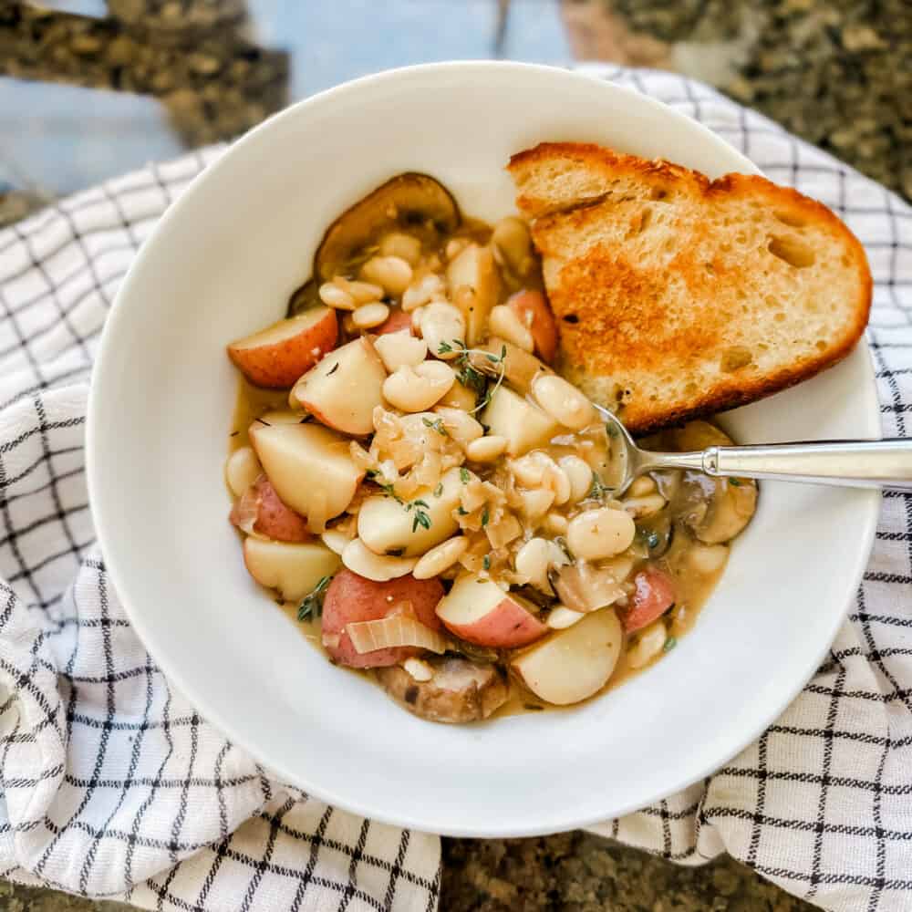 A cozy bowl of Creamy Mushroom and White Bean Stew, featuring a rich, creamy broth filled with tender slices of mushrooms, soft cubes of baby potatoes, and creamy white beans. The stew is garnished with a sprinkle of fresh thyme and rosemary on top, adding a pop of green to the warm, earthy tones of the dish.