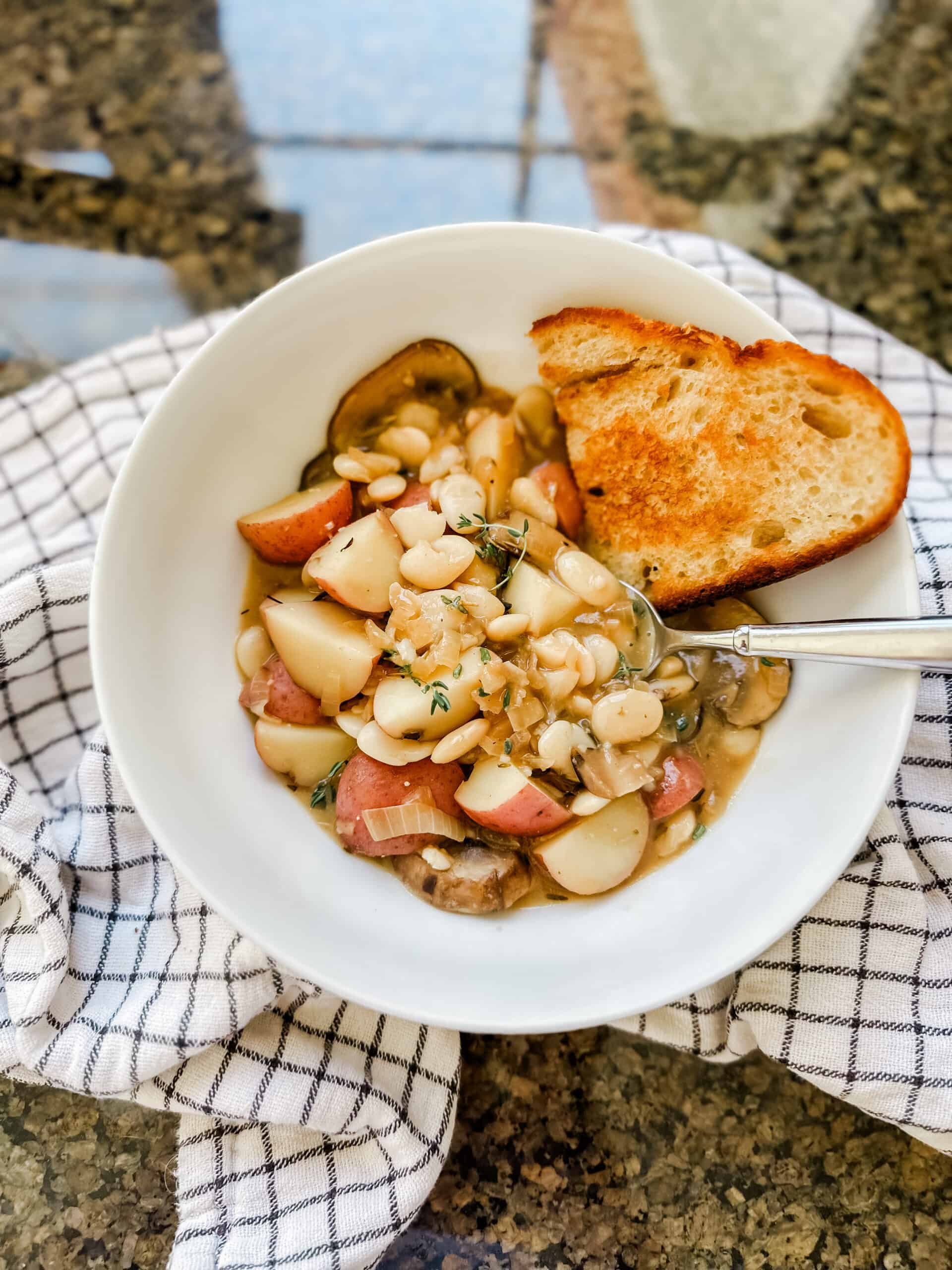 A cozy bowl of Creamy Mushroom and White Bean Stew, featuring a rich, creamy broth filled with tender slices of mushrooms, soft cubes of baby potatoes, and creamy white beans. The stew is garnished with a sprinkle of fresh thyme and rosemary on top, adding a pop of green to the warm, earthy tones of the dish.